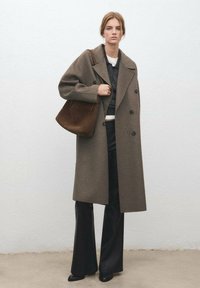 Lange, double-breasted jas van donkerbruin wol, gecombineerd met een donkere trui en zwarte broek. Een bruine suède tas hangt over de schouder.