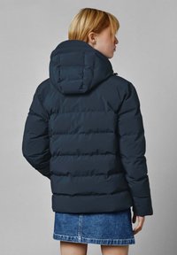 Oseba s kratkimi blond lasmi, oblečena v temno modro puffer jakno z kapuco in kratko denim krilo, obrnjena stran od kamere.
