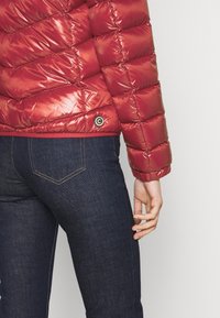 Doudoune rouge avec une finition brillante, présentant des sections matelassées horizontales et un accent de logo à l'ourlet, associée à un jean foncé.