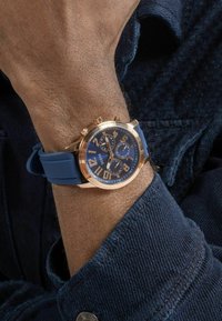Orologio cronografo con cinturino in gomma blu, cassa in metallo color oro rosa, quadrante blu, grandi numeri bianchi e tre sotto-quadranti.
