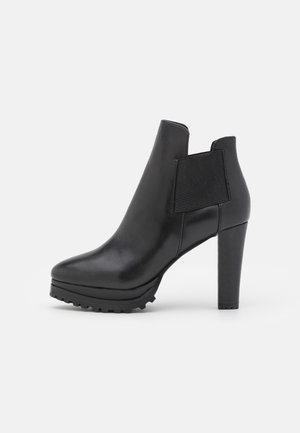 Bota de tobillo negra de cuero con tacón alto cuadrado, suela gruesa de banda de rodadura y panel elástico lateral para facilitar el calce, sobre fondo blanco.
