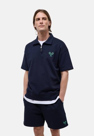 Homme portant un haut marine à demi-fermeture éclair à manches courtes et un short assorti avec des logos verts de raquettes de tennis croisées sur la poitrine et la jambe.