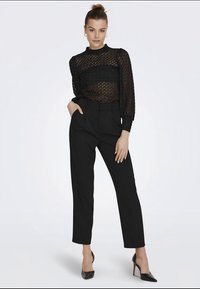 Czarna przezroczysta bluzka z długim rękawem i teksturowanym wzorem, w zestawie z czarnymi spodniami z wysokim stanem oraz czarnymi szpilkami z ostrym noskiem.