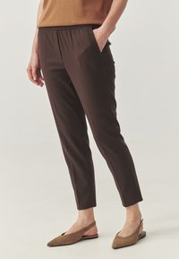 Person, die dunkelbraune, maßgeschneiderte Knöchelhosen mit vorderer Bügelfalte, seitlichen Taschen und hellen spitz zulaufenden Slingback-Sandalen trägt, steht auf einem hellen Boden.