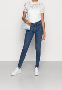 Jeans ajustados de color azul con cintura alta, combinados con una blusa blanca de encaje con patrones florales y un escote redondo. Accesorios con un bolso azul claro.