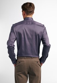 Męska koszula z długim rękawem w ciemnym fiolecie, wykonana z gładkiej tkaniny. Posiada klasyczny kołnierz i mankiety na guziki, zestawiona z khaki spodniami.