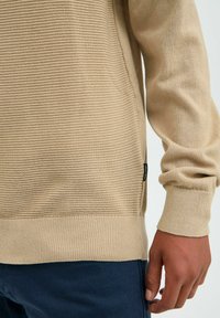 Pull en maille beige avec des rayures horizontales texturées, des poignets et un ourlet côtelés. Étiquette latérale visible ; finition lisse et coupe décontractée.