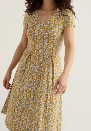 Femme portant une robe jaune moutarde avec un motif floral blanc et bleu clair et des manches courtes, montrée du cou au milieu des cuisses.