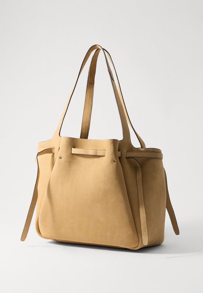 Suède tote bag in licht tan, met twee lange banden, een minimalistisch ontwerp en subtiele stikdetails langs de randen en accenten.
