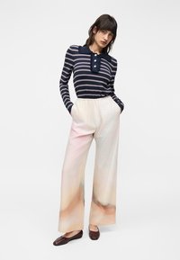 Γυναίκα που φοράει μπλε σκούρο μπλουζάκι με ρίγες και μακριά μανίκια με βολάν στον γιακά και ανοιχτόχρωμο φαρδύ παντελόνι με αφηρημένα παστέλ σχέδια, στέκεται με τα χέρια στις τσέπες.