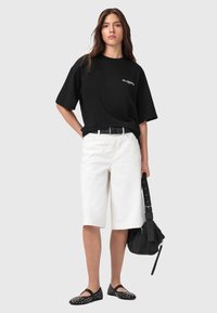 T-shirt nera oversize con lettering bianco, pantaloni corti bianchi fino al ginocchio, cintura nera, ballerine nere con borchie e borsa nera texturizzata.