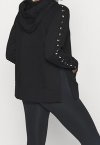 Personne portant un sweat à capuche Nike noir avec un logo le long de la manche et un legging noir, les mains dans les poches du sweat, debout de profil.