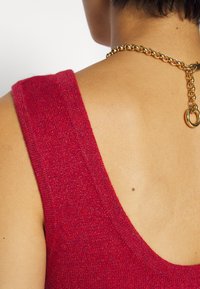 Punane kudumiga tanktop, millel on sügav ümmargune kaelus, tekstureeritud kangas ja kuldne kett detail seljaosas. Kaelal on nähtav sile nahk.
