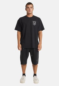 Black cotton t-shirt with short sleeves and a crew neck, featuring a white graphic print on the chest. Worn with black distressed shorts.