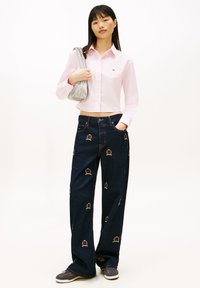 Růžová krátká košile na knoflíky s logem, tmavé široké džíny s barevnou výšivkou a šedá kabelka přes rameno s texturovaným povrchem.