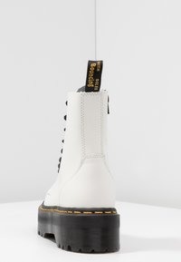 Botín de cuero blanco hasta el tobillo con cordones negros, costuras amarillas y suela de plataforma negra. Cuenta con un tirador trasero con el texto "With Soles".