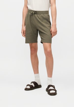 Άτομο που φοράει ελαστικό πλεκτό σορτς χρώματος ελιάς, άσπρες κάλτσες και σκούρα καφέ σανδάλια με πόρπη, στέκεται μπροστά σε απλό φόντο.