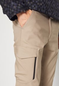 Beige cargobyxor med en slät textur, utrustad med sidofickor och en dragkedjeficka. Bärs med en mörk rutmönstrad topp.