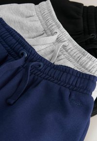Trois pantalons de survêtement à taille élastique soigneusement empilés en bleu marine, gris clair et noir, chacun avec des cordons assortis et un petit logo brodé.
