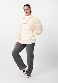 Sudadera de forro polar color crema con media cremallera, bolsillo delantero y tres corazones bordados en tonos pastel centrados en el pecho. Combinada con jeans grises.