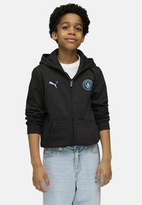 Crna zip-up dukserica s kenguruvim džepom, plavi Puma logo i grb Manchester Cityja; nošena preko bijele majice i svijetlih traperica.