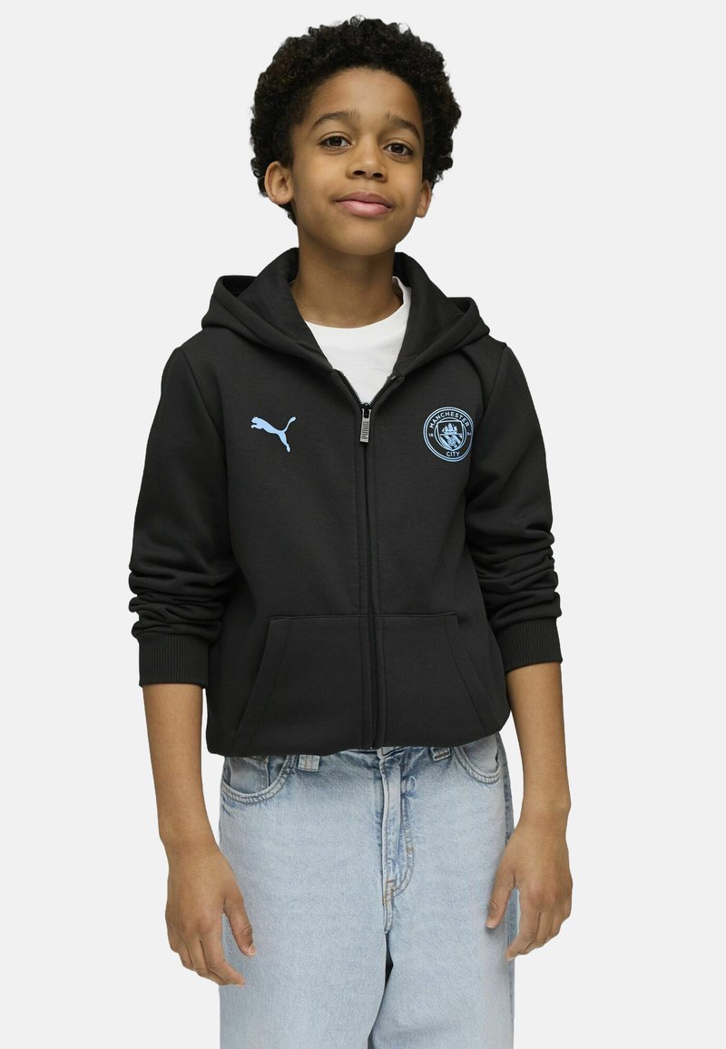Crna zip-up dukserica s kenguruvim džepom, plavi Puma logo i grb Manchester Cityja; nošena preko bijele majice i svijetlih traperica.
