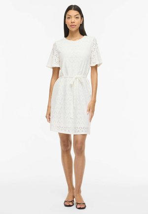 Kvinde iført knælængde hvid øjlet kjole med korte ærmer, bælte i taljen og sorte sandaler med åben tå, stående på hvid baggrund.