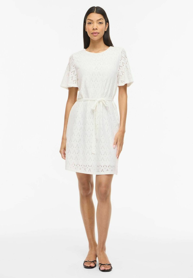 Femme portant une robe blanche à œillets longueur genou, manches courtes, taille ceinturée et sandales noires à bouts ouverts, debout sur fond blanc.