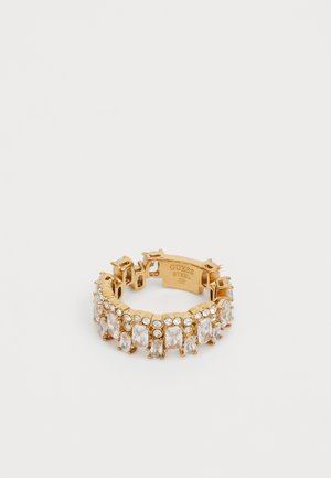 Gold-colored ring featuring a row of clear crystals in various shapes. Made of steel, with engraved brand details on the inner band.