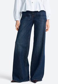 Široké džíny z tmavého denimu, s vysokým pásom, hladkou textúrou a minimálnymi švovými detailmi. Kombinované s bielou blúzkou.