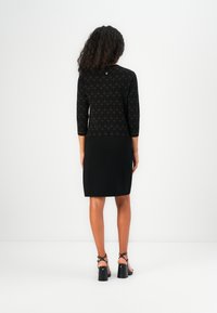 Robe noire avec des manches trois-quarts présentant un motif texturé en haut. Tissu lisse avec une coupe ajustée, se terminant au-dessus du genou. Chaussures à talons hauts.