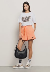 T-shirt grafico bianco con stampa leopardata, pantaloni corti arancioni con coulisse, borsa in pelle nera, sneakers con motivo, calze bianche e nere a righe.