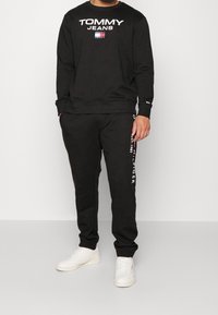 Zwarte katoen sweatshirt met het "TOMMY JEANS" logo, gecombineerd met zwarte sweatpants met witte tekst. Witte sneakers maken de outfit compleet.