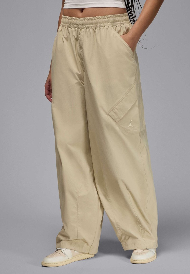 Pantalons beiges amples en tissu léger avec une taille élastique, des poches latérales et un logo de marque discret sur la cuisse.