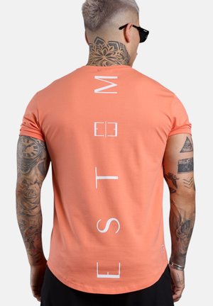 Mann mit Tattoos, der ein korallenrotes T-Shirt mit weißem vertikalem "ESTM"-Schriftzug auf dem Rücken, schwarze Sonnenbrille und schwarze Hose trägt, mit dem Rücken zur Kamera steht.