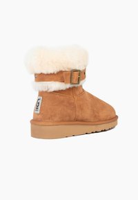 Braune Wildleder-Ankle-Boots mit einem cremefarbenen Kunstfellbund, seitlichem Schnallenverschluss und einer strukturierten Gummisohle. Silbernes Logo-Patch auf der Rückseite.