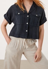 Camicia corta navy in tessuto leggero, con due tasche sul petto, maniche corte arrotolate e bottoni bianchi. Abbinata a pantaloni in lino beige.