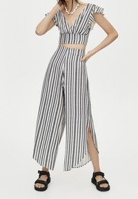 Gestreepte crop top met rimpels mouwen, gecombineerd met brede pantalons met hoge split. Materiaal lijkt lichtgewicht in grijs en witte strepen. Zwarte sandalen.