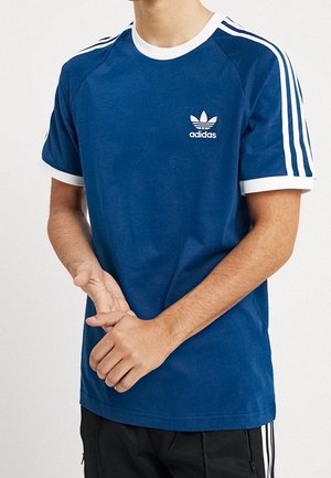Personne portant un t-shirt bleu Adidas avec des bandes blanches sur les manches et un logo sur la poitrine, associé à un pantalon noir avec des bandes blanches.