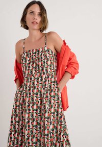 Blommönstrad ärmlös klänning i kräm, grön och rosa, med fyrkantig halsringning och fickor, kombinerad med en klar röd jacka.