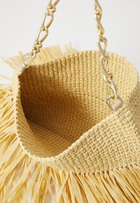 Sac en paille tissé avec un haut ouvert et effilé. Il est doté d'une bandoulière en chaîne dorée et d'une texture frangée sur le bas. Couleur : jaune clair.