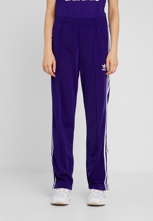 Osoba w fioletowych spodniach dresowych Adidas z białymi paskami po bokach i białych butach sportowych, stojąca na jednolitym tle.