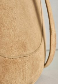 Puha, barackszínű velúr táska sima textúrával, ívelt sziluettel és egyetlen puha pánttal. A varrási részletek láthatók a felületen.