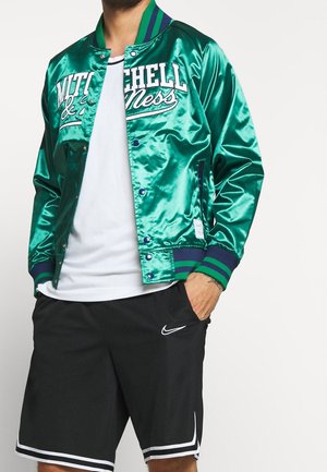 Man draagt een groene satijnen Mitchell & Ness varsity jas, wit overhemd, zwarte Nike short met witte rand, staat met één hand in de zak.