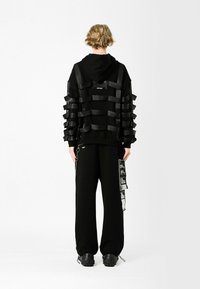 Sudadera negra con diseño de cintas en capas, que cuenta con una capucha grande y una etiqueta. Combinada con pantalones sueltos negros y zapatos gruesos negros.