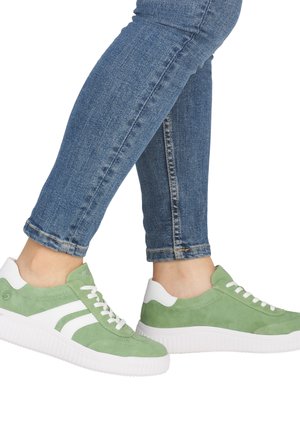 Person wearing blue jeans and light green and white low-top sneakers standing with one foot slightly raised.