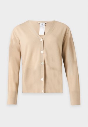 Femme debout portant un cardigan beige boutonné, un pantalon large gris clair et des chaussures noires pointues sur un fond clair uni.