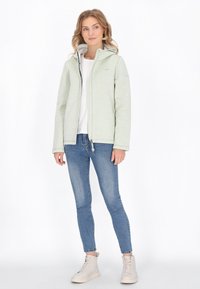 Jasnozielona kurtka zapinana na zamek z kapturem, o gładkiej fakturze i kontrastowym wnętrzu. W połączeniu z dopasowanymi dżinsami i beżowymi butami.