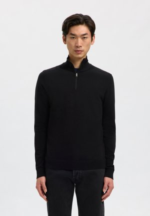 Man draagt een zwarte trui met lange mouwen en een halve ritssluiting bij de kraag en een donkere broek, staand tegen een effen lichte achtergrond.