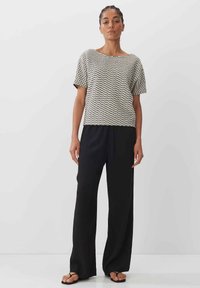 Un haut à manches courtes au motif en zigzag avec des couleurs crème et noir, associé à un pantalon noir ample et des sandales plates, se tenant contre un fond blanc.